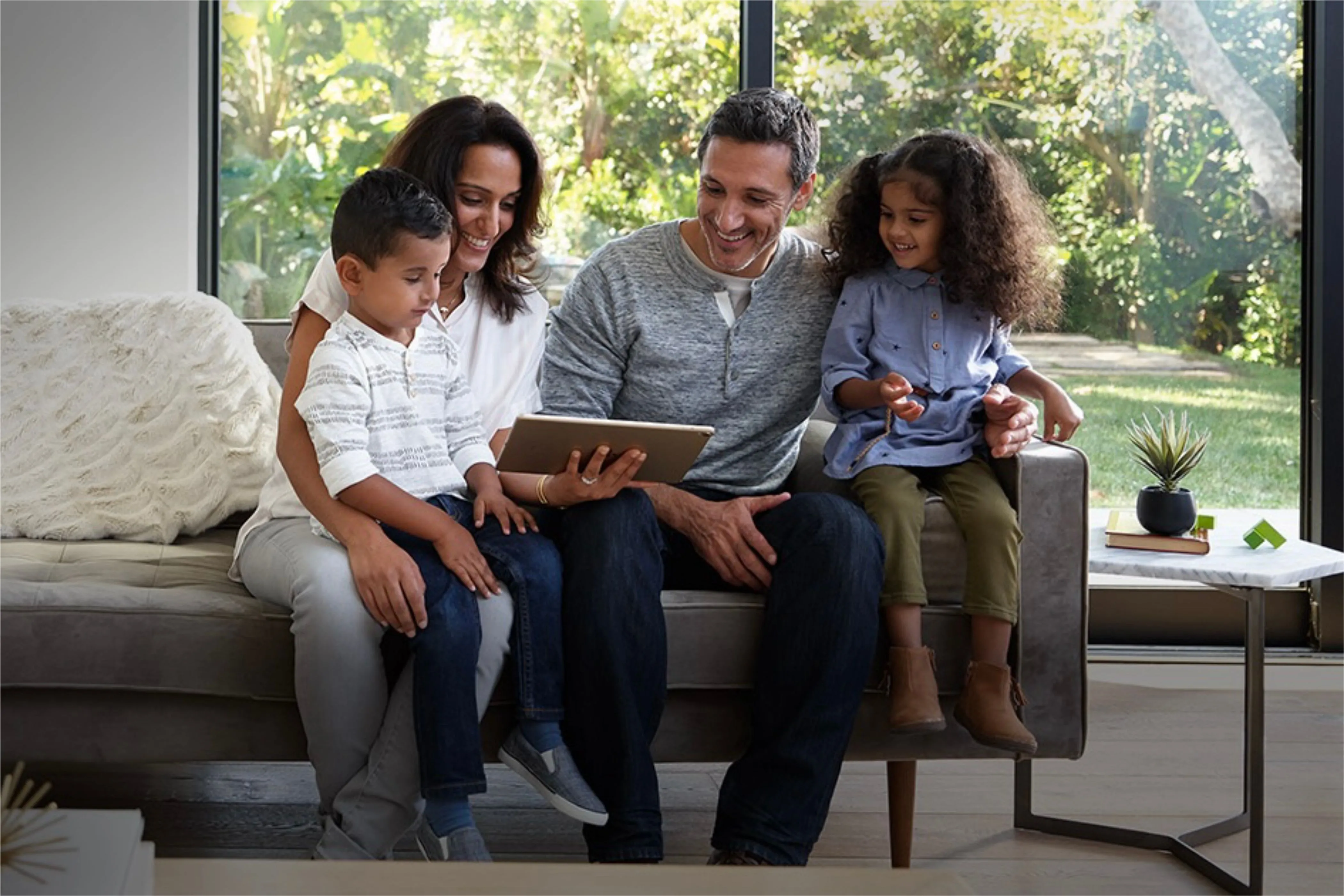 Hai người lớn và hai trẻ em tập trung quanh một chiếc iPad trên ghế sofa, minh họa tính năng Family Sharing.