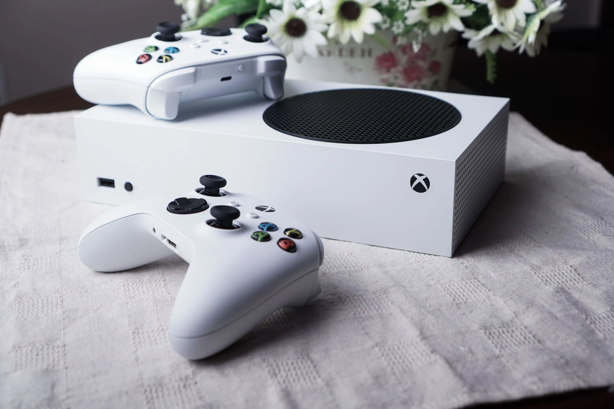 Máy chơi game Xbox Series S đặt trên bàn cùng hai tay cầm điều khiển và một lọ hoa, thể hiện sự nhỏ gọn và vai trò của nó trong phân khúc console giá mềm.