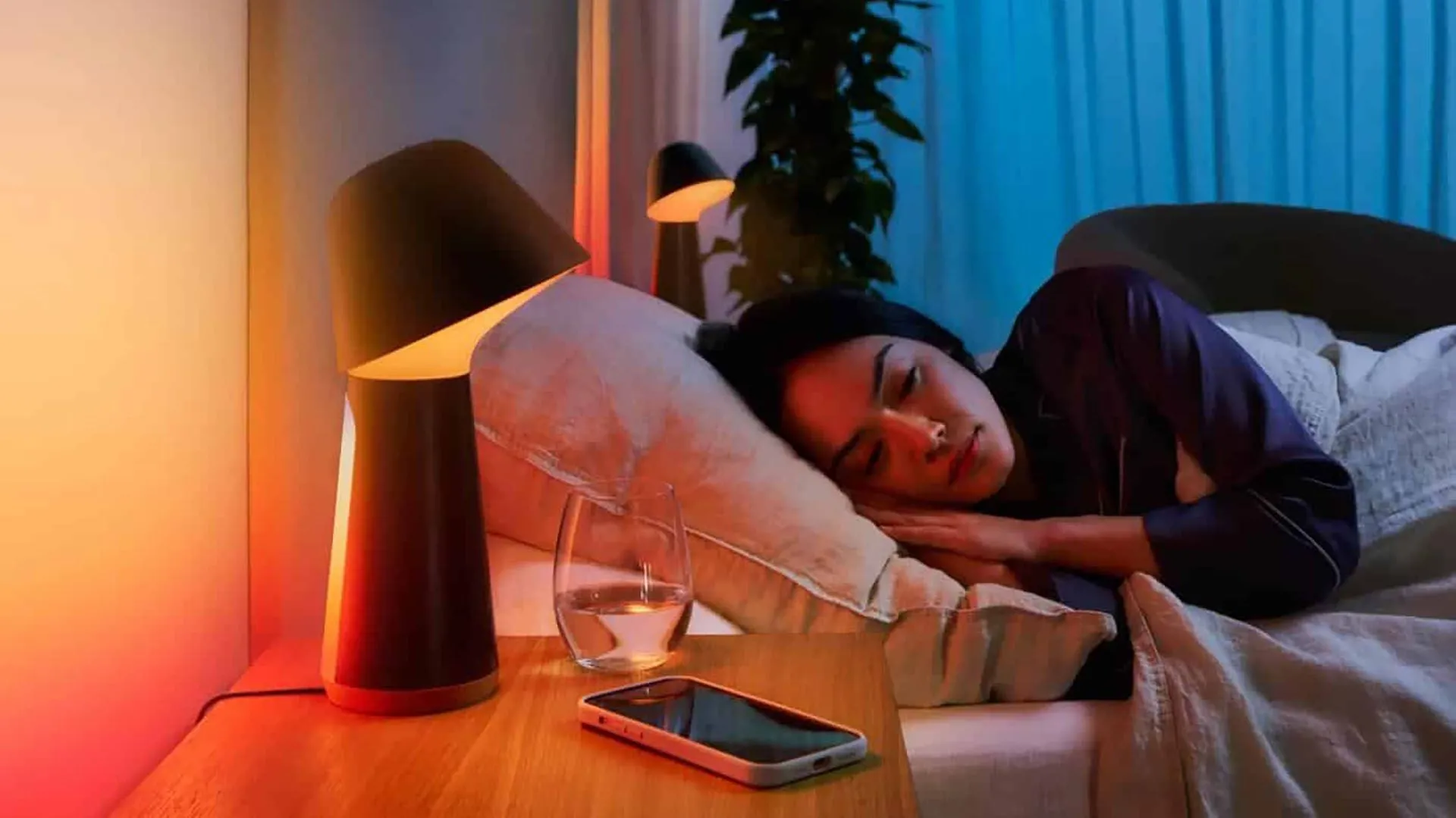 Người phụ nữ ngủ ngon lành trên giường, gần đó có đèn thông minh và điện thoại thông minh đang phát tiếng ồn trắng hỗ trợ giấc ngủ.