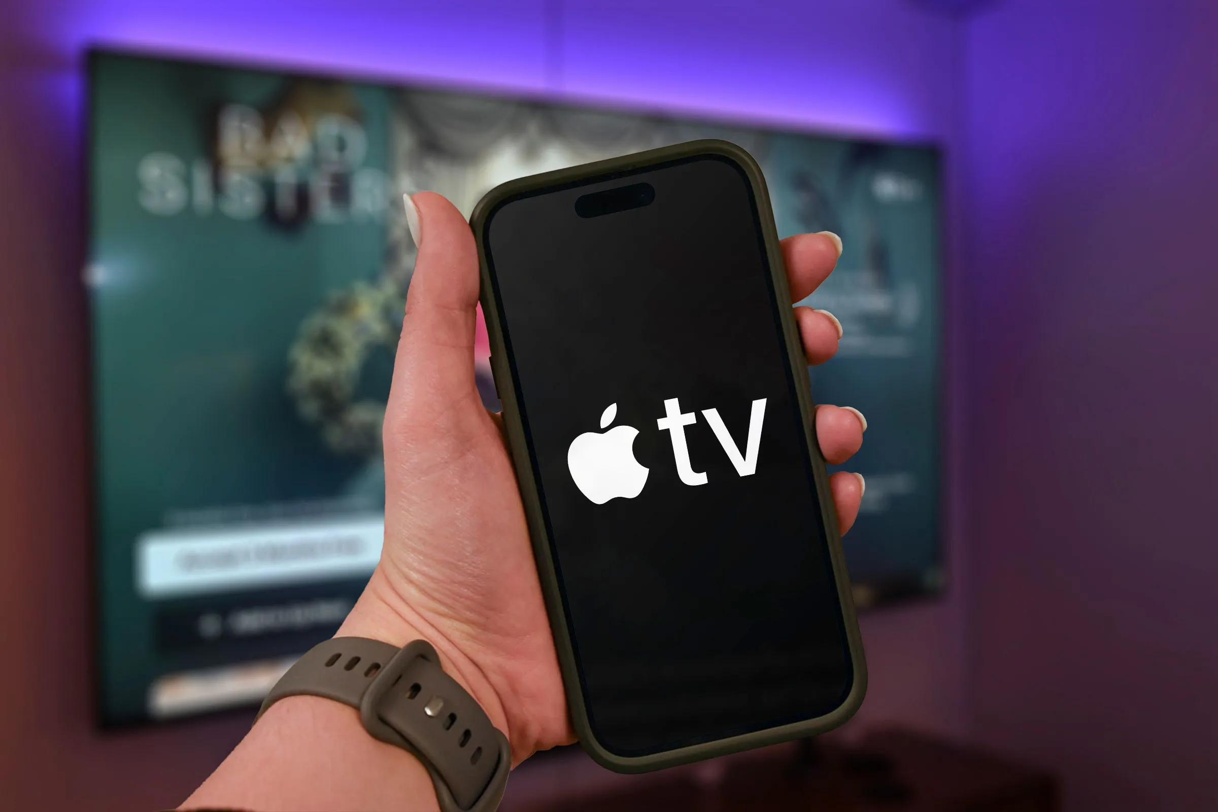 Bàn tay cầm iPhone hiển thị logo Apple TV+ với TV mờ ở hậu cảnh.