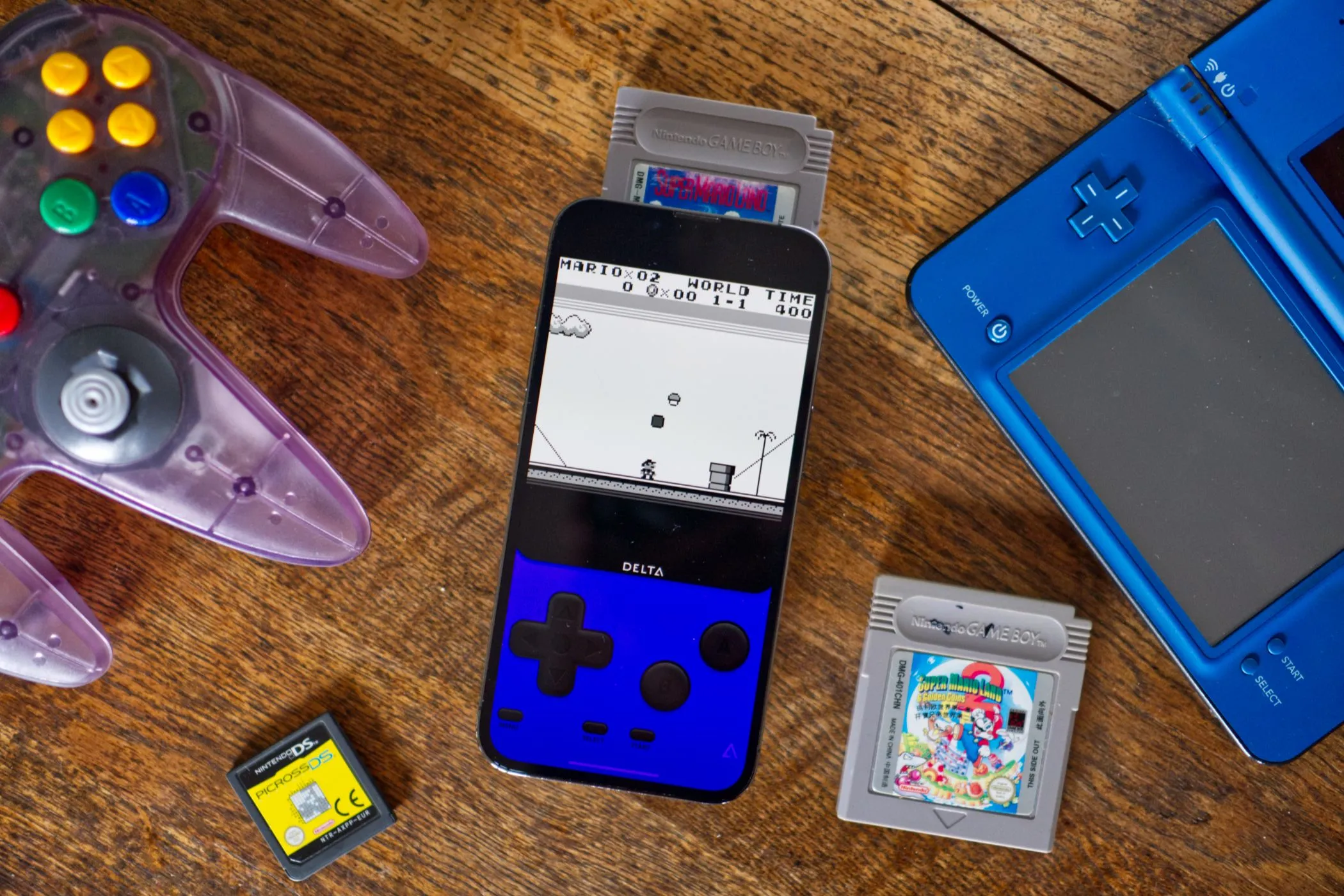 Điện thoại iPhone hiển thị ứng dụng giả lập Delta, đặt cạnh bộ điều khiển Nintendo 64, máy Nintendo DSi và nhiều hộp băng game cổ điển.